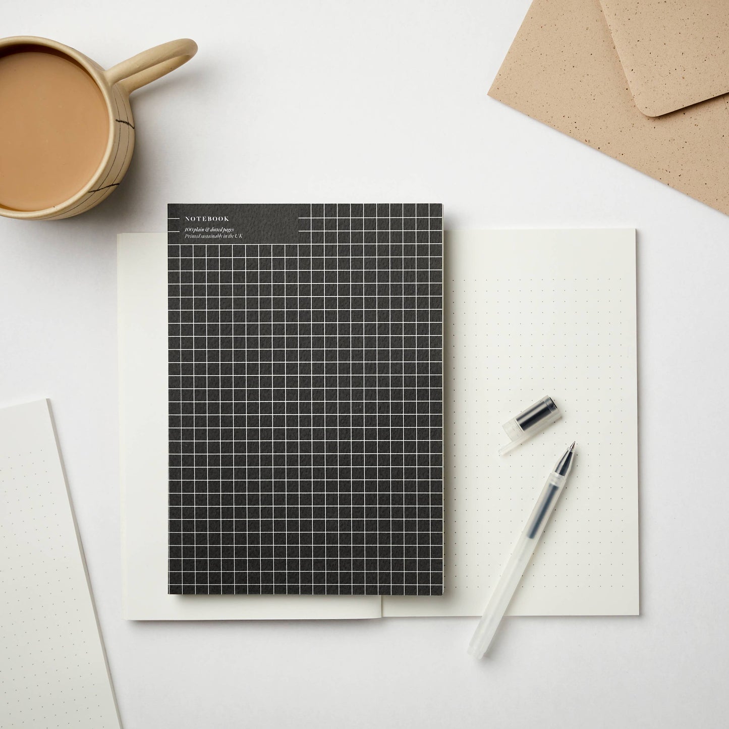 Black A5 sketchbook with white grid cover, placed on open dotted notebook with pen and coffee cup – ideal for creatives and journaling.