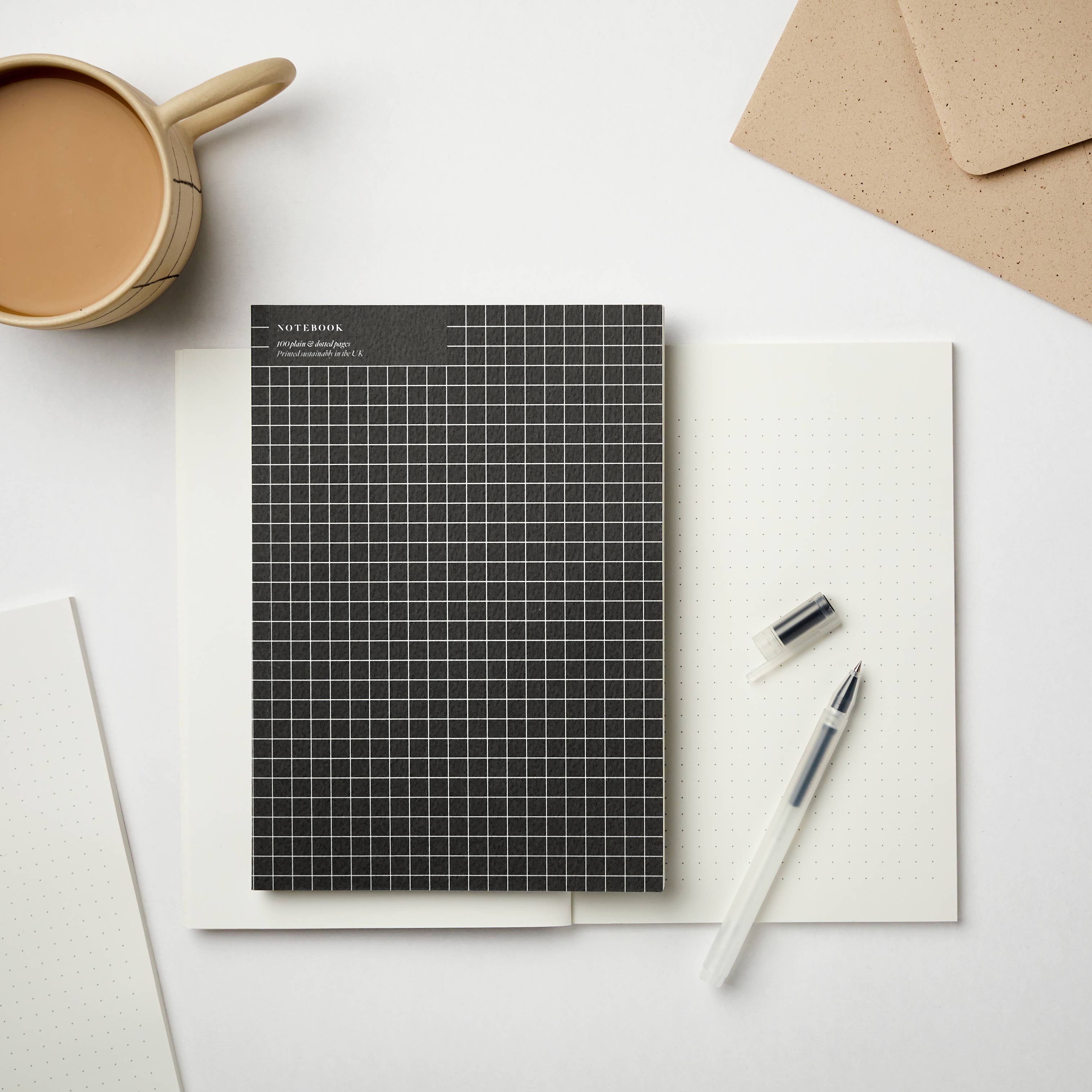 Black A5 sketchbook with white grid cover, placed on open dotted notebook with pen and coffee cup – ideal for creatives and journaling.